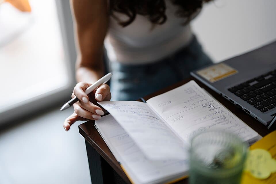 Personne prenant des notes au stylo dans un carnet ouvert, posé sur un bureau à côté d'un ordinateur portable